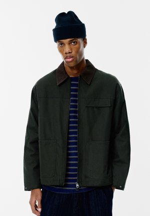 Giacca verde scuro con colletto marrone, dotata di due tasche frontali. Il modello indossa una camicia a righe blu navy e un berretto blu navy.