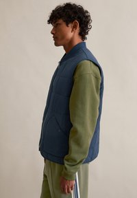 Marineblau gesteppte Weste mit Fronttaschen und Reißverschluss, getragen über einem grünen Langarmshirt, vor neutralem Hintergrund präsentiert.