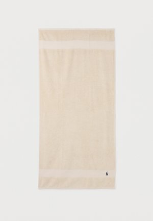 Serviette en coton beige avec deux bandes horizontales près des bords et un petit logo brodé bleu foncé dans le coin inférieur droit.