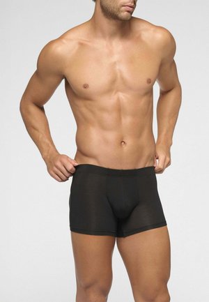 Uomo senza maglietta con corporatura muscolosa, indossa boxer neri e sistema la cintura davanti a uno sfondo chiaro uniforme.