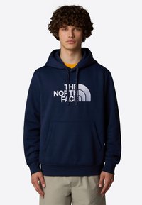 Sudadera azul marino con un bolsillo tipo canguro, capucha con cordón y un gran logo blanco que dice "THE NORTH FACE" en el frente. Textura suave.