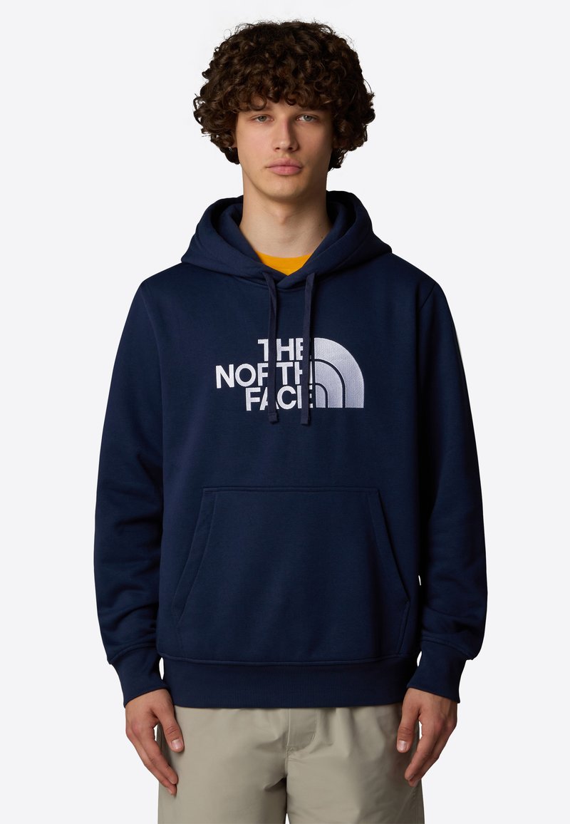 Sudadera azul marino con un bolsillo tipo canguro, capucha con cordón y un gran logo blanco que dice "THE NORTH FACE" en el frente. Textura suave.