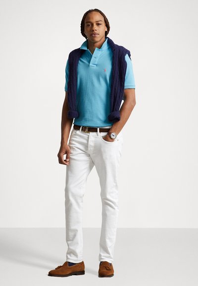 Tricou polo de culoare bleu deschis cu guler, pulover închis la culoare aruncat pe umeri, pantaloni albi și pantofi din piele ante maro. Ceas argintiu la încheietura mâinii.