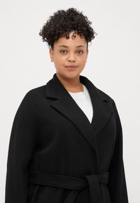 Mustat vyötäröllä varustettu takki, jossa on kaulus ja teksturoitu kangas. Muotoilussa on kietaisu-tyyli ja taskut. Päälle puettuna valkoinen paita.