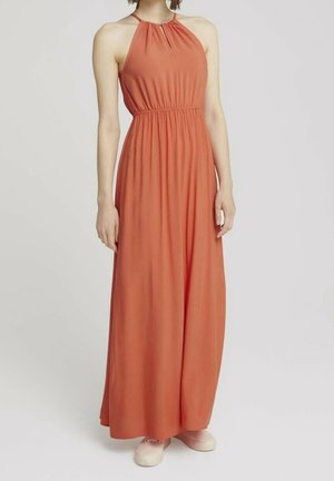Femme portant une robe maxi sans manches couleur rouille avec un col halter et une taille froncée, associée à des baskets rose clair, debout sur un fond uni.