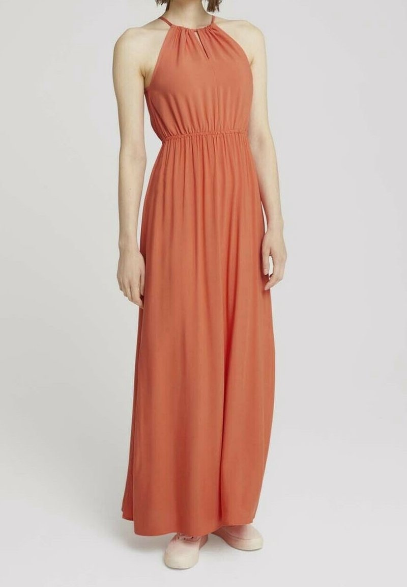Femme portant une robe maxi sans manches couleur rouille avec un col halter et une taille froncée, associée à des baskets rose clair, debout sur un fond uni.