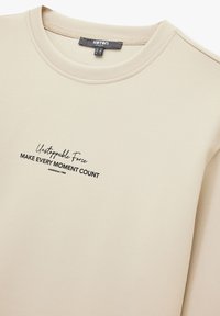 Beige sweatshirt med rundad halsringning, med en svart tryckt textdesign: "Ostoppbar kraft GÖR VARJE ÖGONBLICK VÄRDEFULLT" och etableringsår.