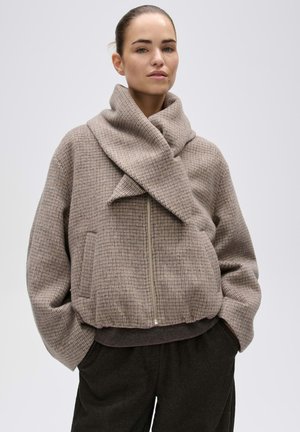 Vrouw draagt een beige geruit jack met rits en een grote overslagkraag en donkere broek, staand met handen in de zakken.