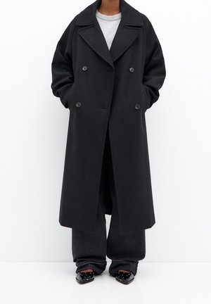 Personne portant un long manteau noir croisé, un haut gris, un pantalon large noir et des chaussures pointues noires à pois blancs sur fond blanc.