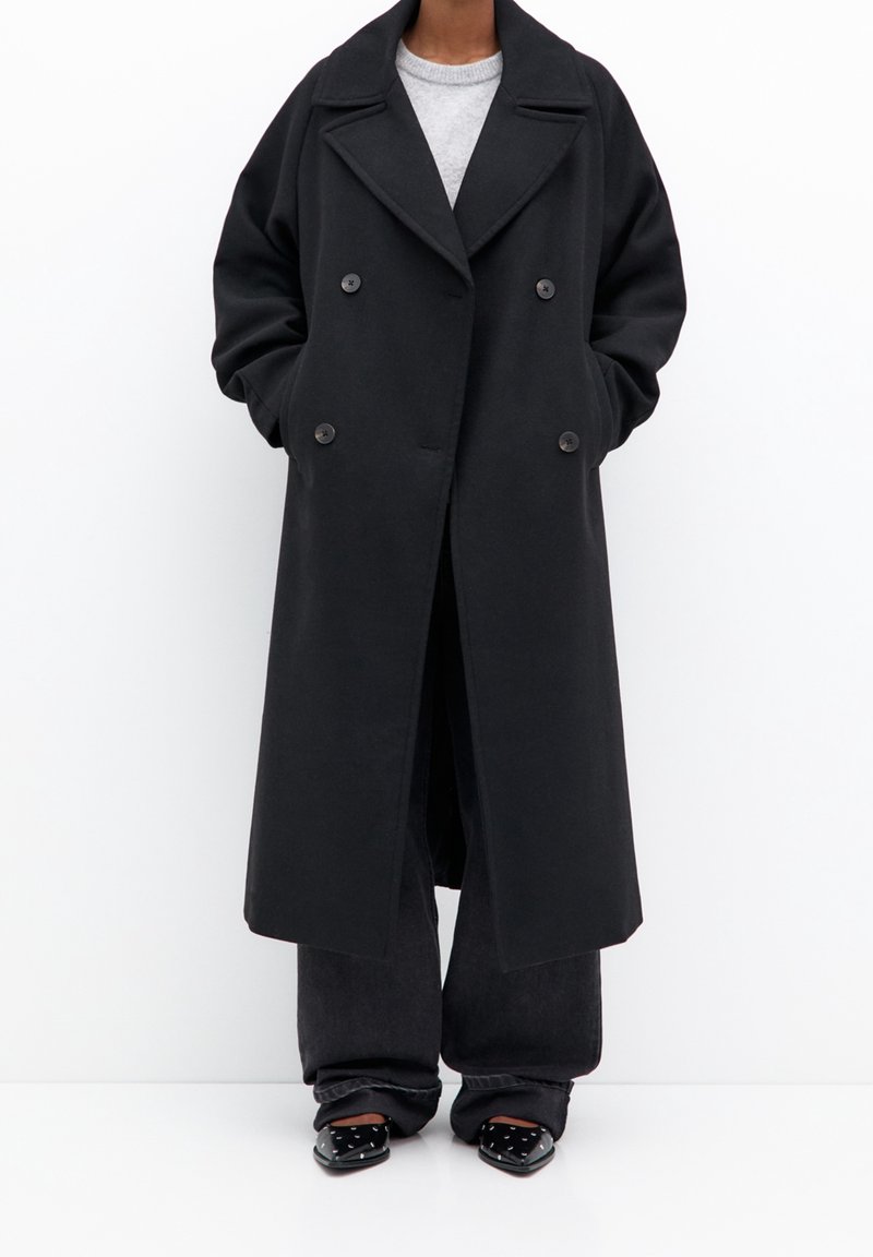 Personne portant un long manteau noir croisé, un haut gris, un pantalon large noir et des chaussures pointues noires à pois blancs sur fond blanc.