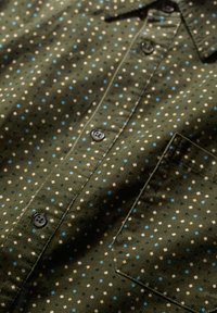 Camicia verde a fantasia con piccoli punti bianchi, blu e gialli. Realizzata in tessuto con una finitura texturizzata, include bottoni neri e una tasca sul petto.