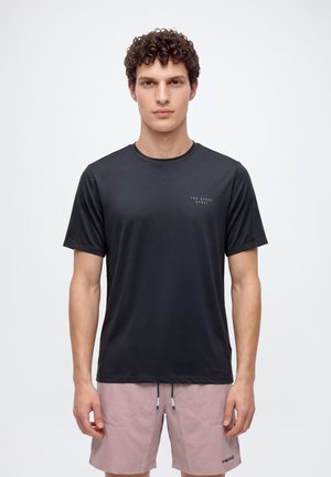 Jeune homme aux cheveux bouclés portant un t-shirt noir Ted Baker Sport et un short HEAD rose clair, debout devant un fond blanc uni.