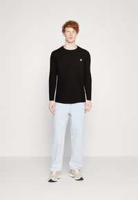 Svart langermet t-skjorte med en liten rød trekantet logo på venstre bryst, kombinert med lyseblå jeans og beige sneakers.
