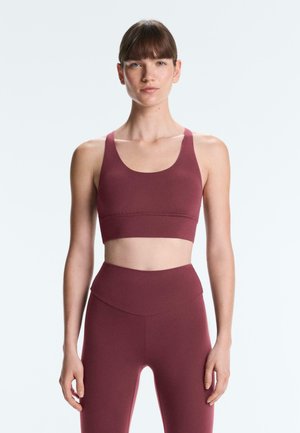 Tânără stând cu brațele relaxate, purtând un sutien sport și colanți cu talie înaltă, ambele de culoare burgundy, pe un fundal simplu și luminos.