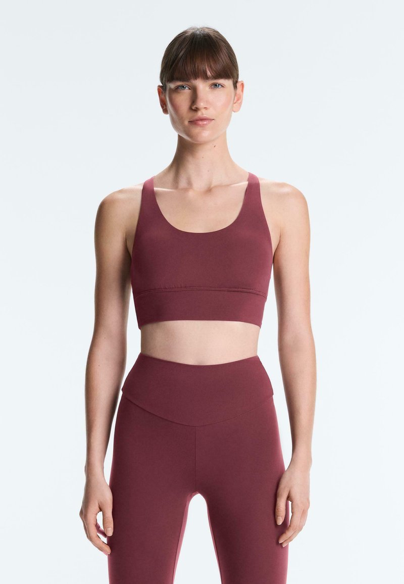 Fiatal nő áll lazán lógó karokkal, bordó sportmelltartót és magasderekú leggingset viselve, egyszínű világos háttér előtt.