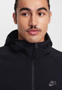 Chaqueta negra con cremallera y capucha, con costuras en contraste y un logo reflectante de Nike en la parte superior izquierda, hecha de material ligero.