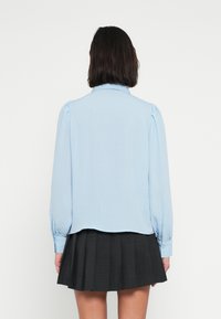 Γαλάζιο μπλουζάκι με μακριά, φουσκωτά μανίκια, γιακά και στρογγυλεμένο τελείωμα. Φορεμένο με μαύρη πλισέ φούστα. Ομαλή υφή.