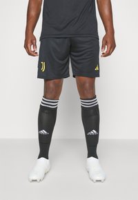 Pantalones cortos deportivos negros con el logo amarillo de Juventus, combinados con calcetas negras hasta la rodilla que tienen la marca blanca de Adidas y zapatillas blancas.