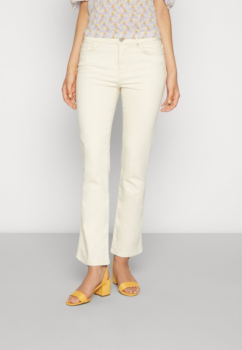 Vaqueros acampanados color crema con una textura suave. Combinados con una blusa estampada y sandalias amarillas de tacón cuadrado. Corte de pierna recta.