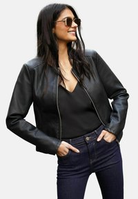 Black leather jacket with a cropped design, silver zipper, and shoulder seams. Worn over a black top and dark denim jeans. Sunglasses included.