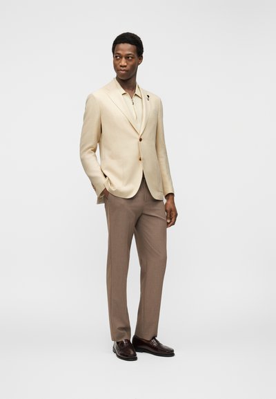 Άνδρας που στέκεται με τα χέρια στις τσέπες, φορώντας μπεζ σακάκι, ανοιχτό μπεζ πουκάμισο, καφέ παντελόνι και σκούρα καφέ παπούτσια.