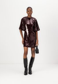 Robe en sequins bordeaux avec une silhouette droite et des manches évasées, associée à des bottes noires montantes et un petit sac à main noir.