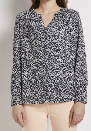 Vrouw die een zwarte blouse met lange mouwen met wit luipaardprintpatroon en lichtbeige broek draagt tegen een effen achtergrond.