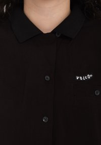 Chemise noire avec un tissu texturé, dotée d'un col noir, d'une fermeture à boutons et d'un patch de logo blanc VOLCOM sur la poche gauche de la poitrine.