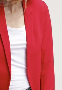 Blazer rouge à texture lisse, coupe ajustée et revers crantés, porté par-dessus un t-shirt blanc. La veste présente des coutures visibles minimales.