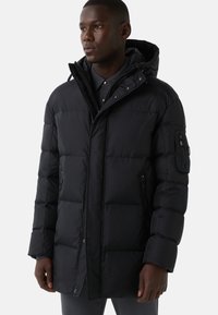 Schwarze Pufferjacke mit Kapuze, ausgestattet mit einem Reißverschluss, seitlichen Taschen und einer strukturierten Oberfläche. Enthält eine aufgesetzte Tasche am Arm.