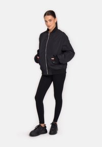 Sort bomberjakke med lynlås, elastiske manchetter og sidelommer, parret med sorte leggings og kraftige sorte sneakers.
