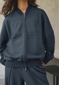 Donkerblauwe bomberjack met rits, gemaakt van zachte stof, met een geribde kraag, grote zakken en een relaxte pasvorm, gecombineerd met bijpassende broek.