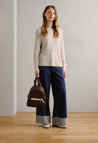Zaino in pelle marrone con forma arrotondata e manico superiore, abbinato a un maglione beige a coste e jeans in denim scuro a gamba ampia con orli a contrasto.