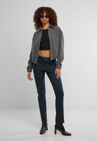 Chaqueta bomber gris con cierre de cremallera, top corto negro ajustado, jeans skinny azul marino y botas negras até. La modelo tiene el cabello rizado y lleva gafas de sol.