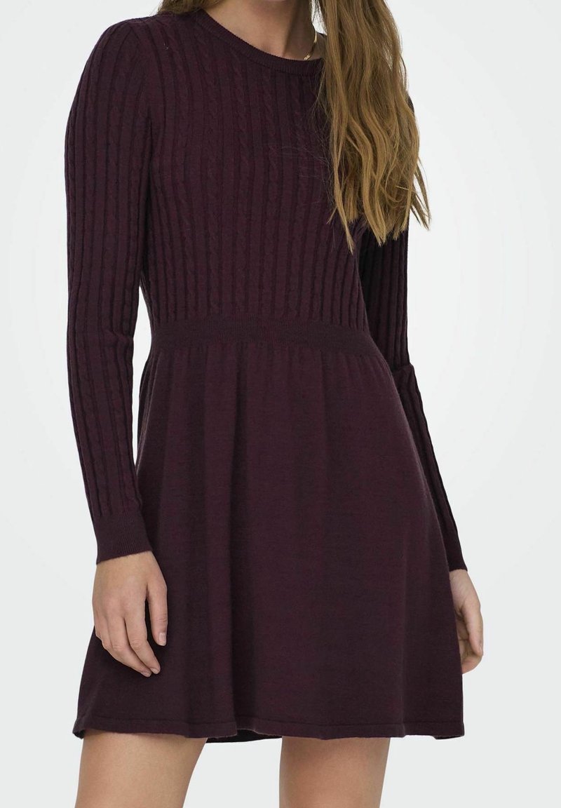 Femme portant une robe en maille à manches longues de couleur violet foncé, avec un corsage côtelé ajusté et une jupe évasée, debout devant un fond uni.