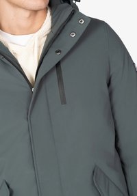 Hombre que lleva una chaqueta con capucha gris oscuro sobre un suéter de punto color crema con detalles visibles de cremallera y botones de presión.