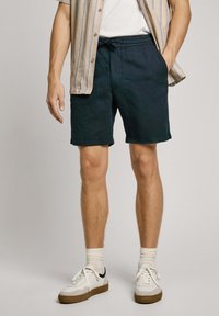 Pantalones cortos verde oscuro de corte holgado, con cinturilla ajustable, bolsillos laterales y dobladillos acabados, combinados con una camisa de rayas de manga corta y zapatillas blancas.