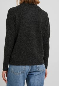 Suéter gris oscuro, texturizado, con un corte relajado, mangas largas y un cuello alto, combinado con jeans holgados de color azul claro.