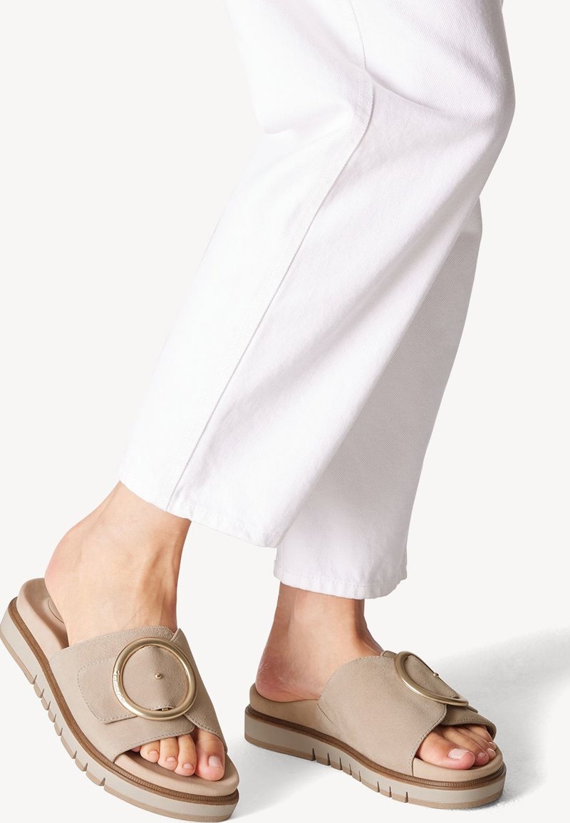 Pieds portant des sandales beige à enfiler avec une grande boucle ronde, associées à un pantalon blanc coupé au-dessus de la cheville sur un fond uni.
