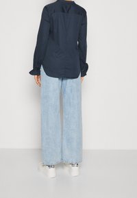 Camisa de manga larga en marino con puños con volantes y textura suave, combinada con pantalones vaqueros anchos de mezclilla clara y zapatillas blancas con acentos metálicos.