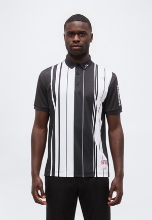 Homme debout face à l’avant, portant un polo à manches courtes rayé noir et blanc et un pantalon noir sur un fond uni.