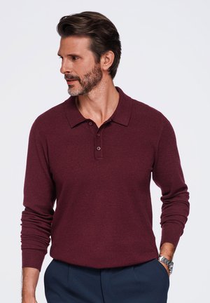 Homme barbu portant un polo bordeaux à manches longues et un pantalon bleu marine, debout, une main dans la poche, regardant vers la gauche sur un fond uni.
