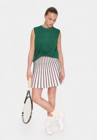 Haut en tricot sans manches vert avec des bordures blanches, associé à une jupe plissée rose avec des rayures verticales vertes. Tenant une raquette de tennis.
