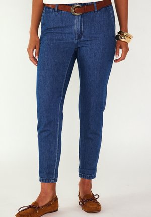 Donna che indossa jeans blu a vita alta con cintura marrone, mocassini marroni e braccialetti dorati al polso destro.