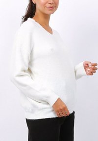 Femme portant un pull en maille blanc à col en V avec des manches longues et un pantalon noir, se tenant contre un fond clair uni.