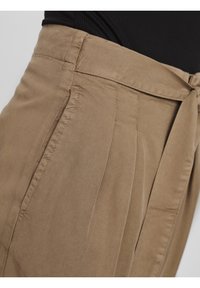 Pantalons taille haute beiges avec une finition texturée, dotés de plis sur le devant et d'un détail de ceinture nouée à la taille, offrant une coupe à la fois structurée et décontractée.