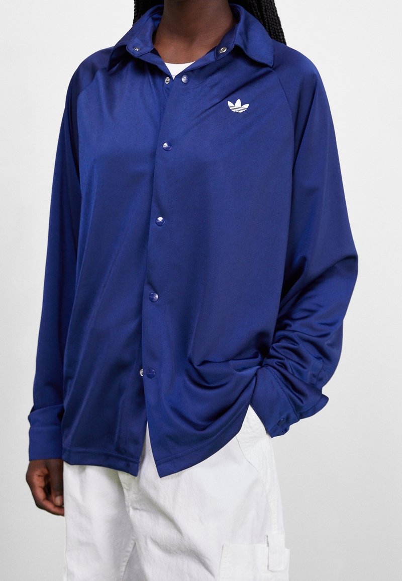 Navy blue, satin button-up jacket with a collar, long sleeves, and subtle white logo on the left chest. Features a relaxed fit.