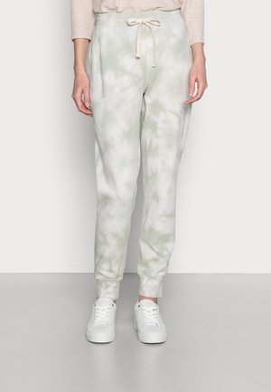 Person iført lysegrønne og hvide tie-dye joggebukser med snøre i taljen og hvide sneakers, stående foran en ensfarvet baggrund.