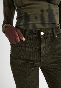 Pantaloni di velluto a coste verde oliva con texture rigata, tasche frontali e chiusura con bottone argento in vita. La mano del modello è appoggiata nella tasca.