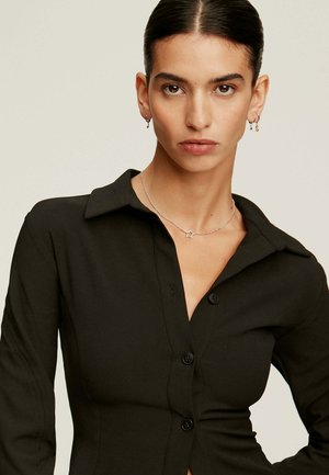 Camisa negra de botones con cuello estructurado, mangas largas y forma entallada; modelo con un collar de estrellas plateadas y pequeños aros en las orejas.